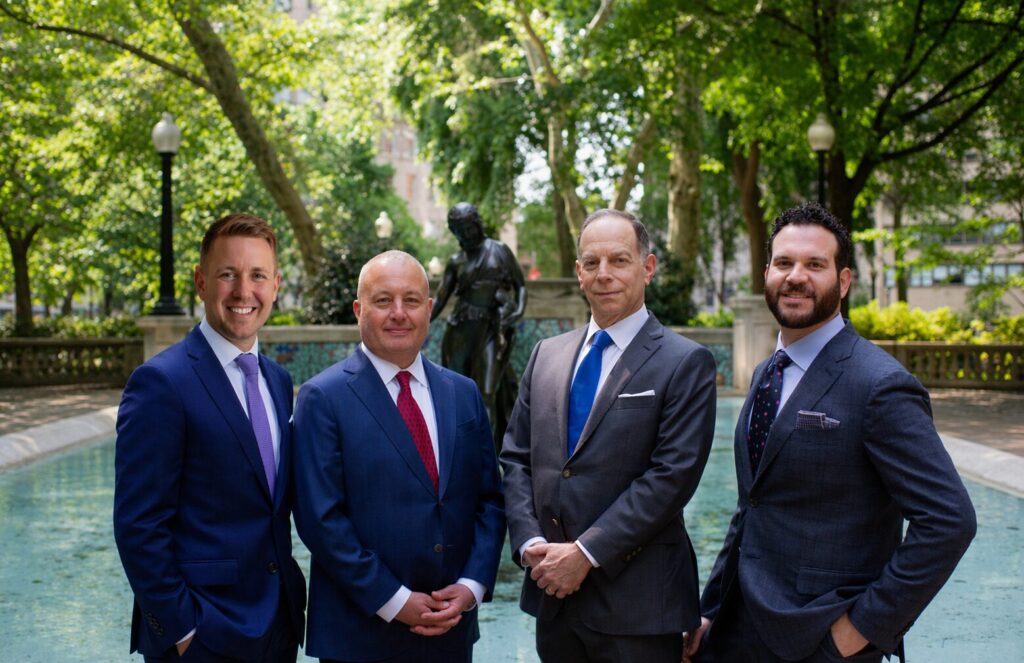 Four businessmen in suits stand side by side near a fountain in a green park.