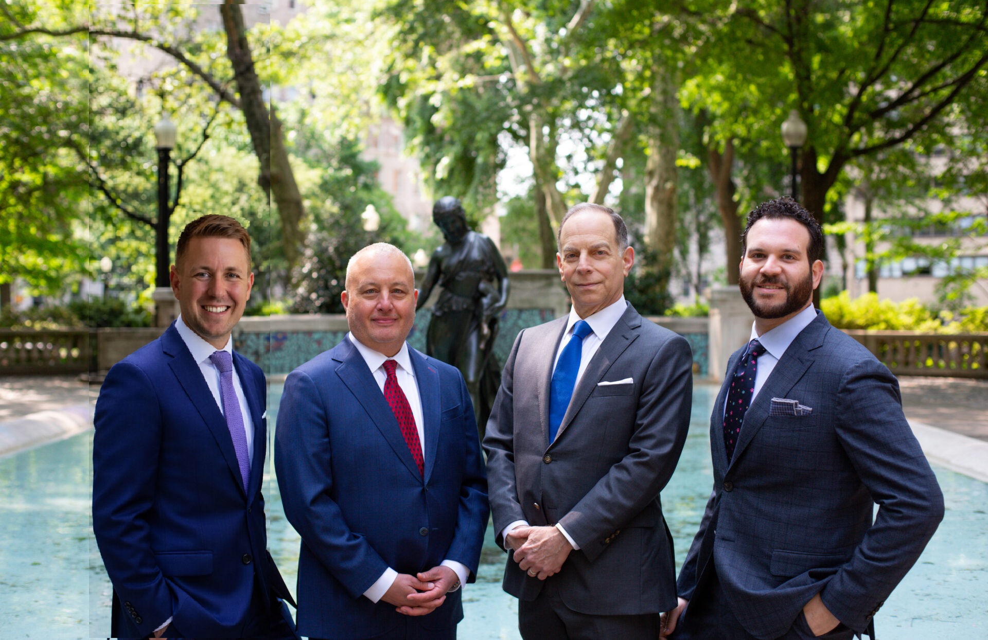 Four businessmen in suits stand together near a fountain in a leafy city park behind them.